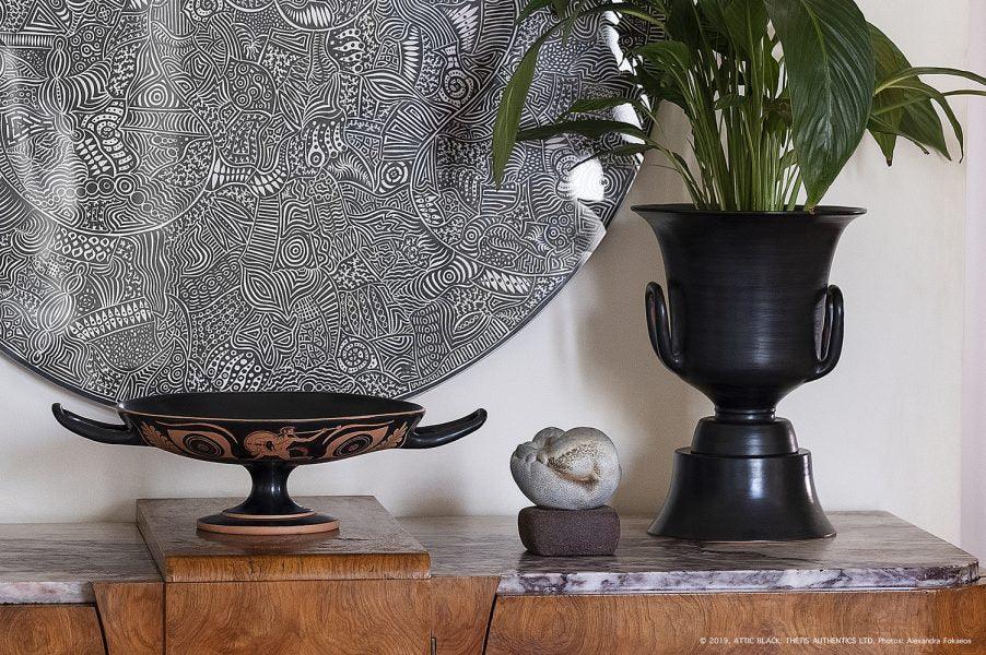 Handcrafted black ceramic tableware, vase with plant, and patterned plate on wooden sideboard.