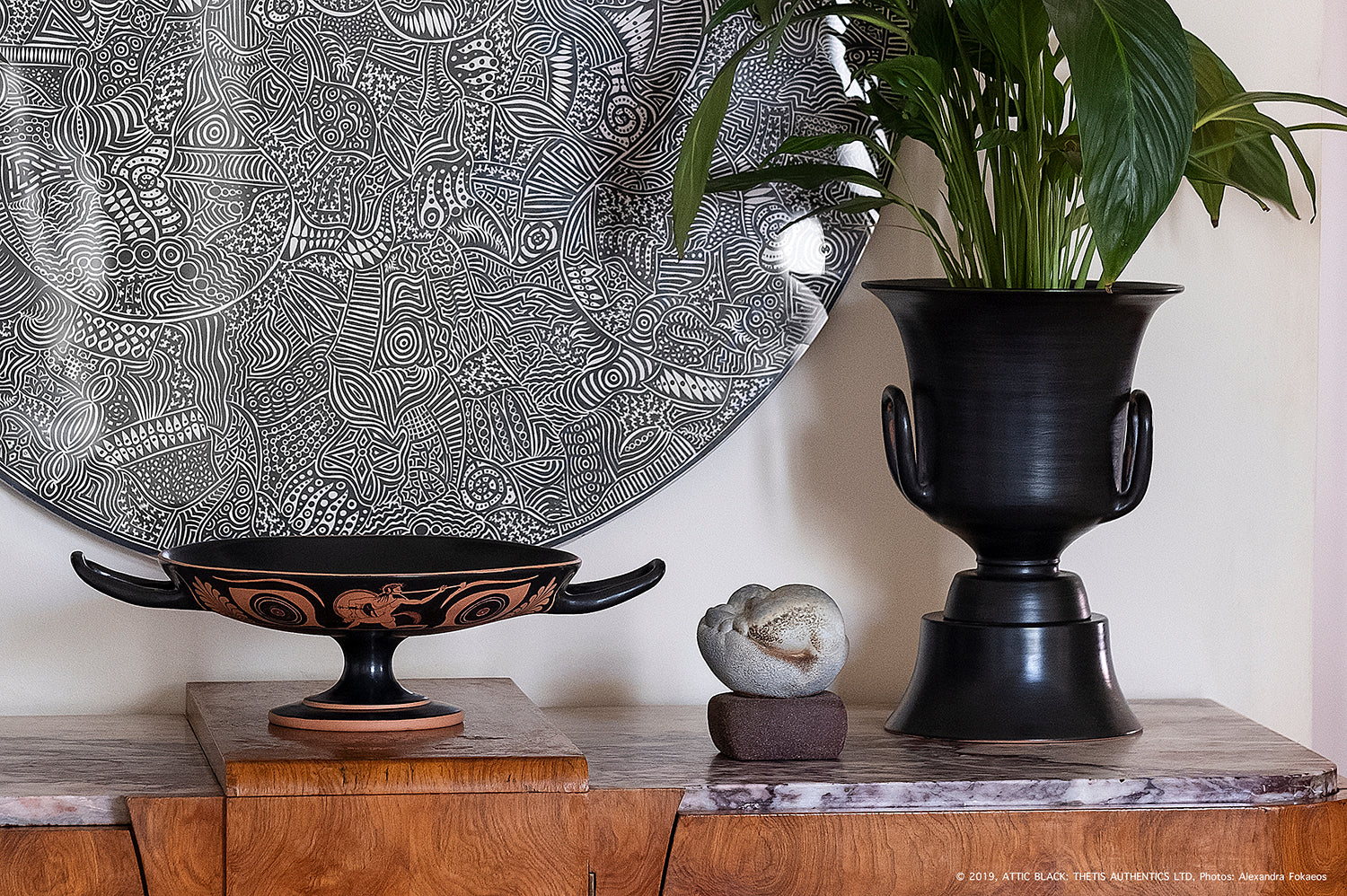 Handcrafted black ceramic vase with plant, ancient Greek kylix, and decorative plate on wooden shelf