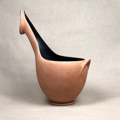 A sculptural ceramic pitcher with a curved neck and spout, made of terracotta-colored clay with a glossy black interior, set against a neutral light gray background.
