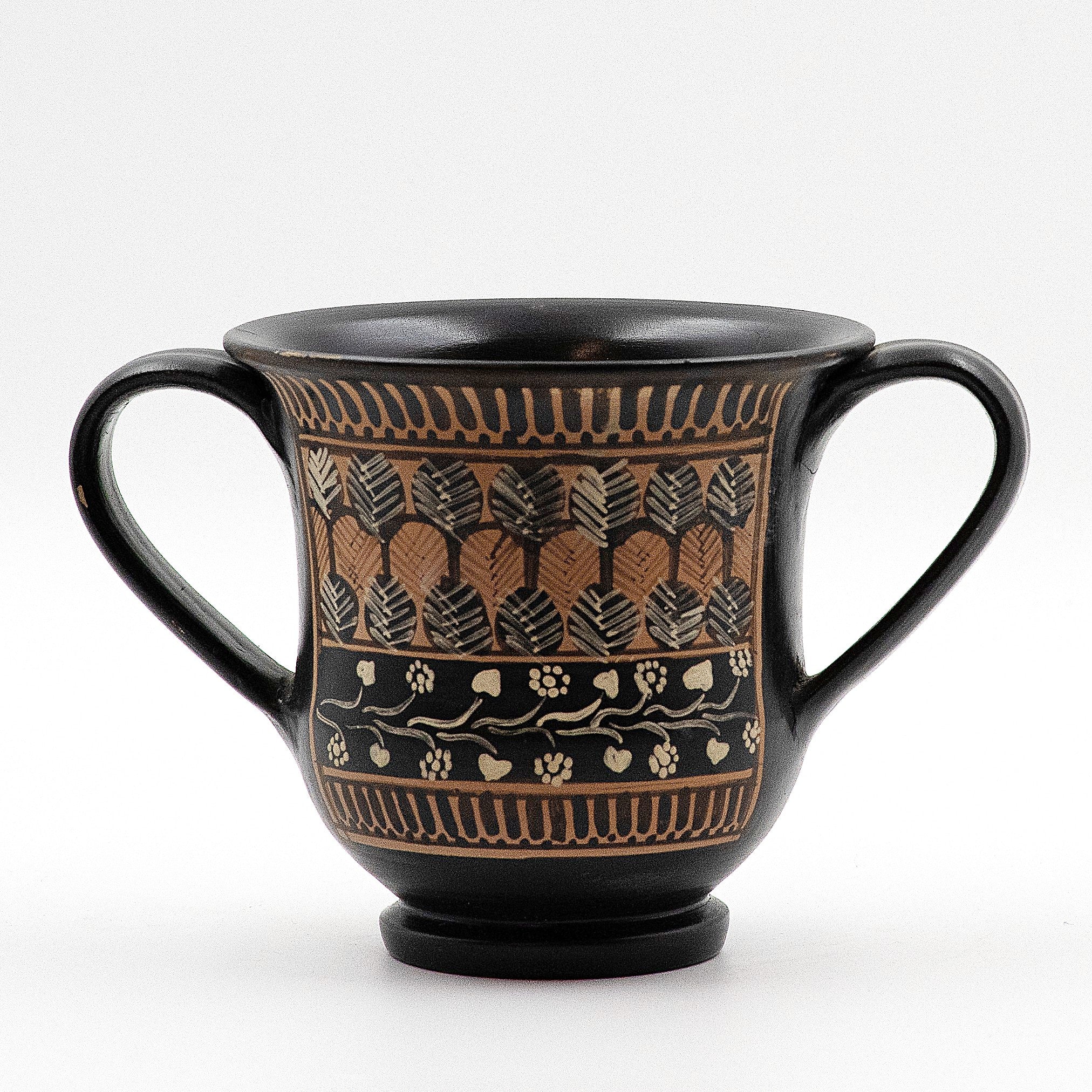 A black ceramic drinking cup with two handles, featuring a stylized foliate pattern and other decorative motifs.