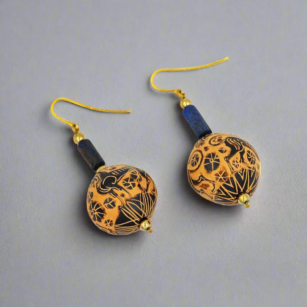 A pair of black-figure drop earrings featuring intricately decorated ceramic beads with gold accents and old lapis elements, displayed against a grey background.