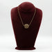 A ceramic pendant necklace displayed on a red velvet mannequin bust. The necklace has a gold-colored chain and a brown, circular pendant.