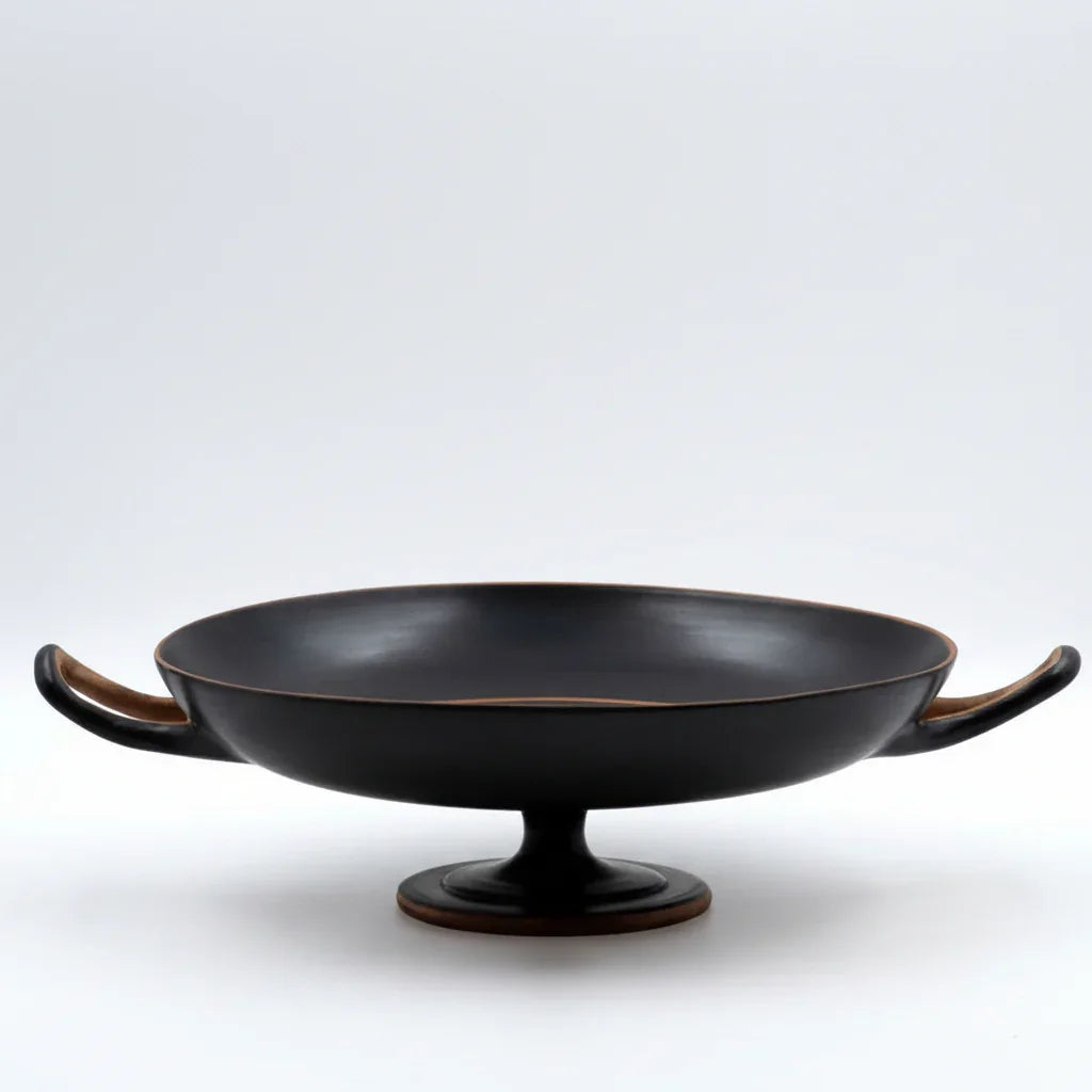 Hand-thrown black ceramic kylix bowl with two handles and pedestal base on white background