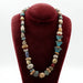 A Mycenaean bead necklace displayed on a red stand, featuring a variety of blue and multicolored beads made from faience, ceramic, and semi-precious stones, with a gold-plated silver clasp.