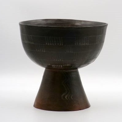 This image features a striking round pedestal vase, distinctively crafted with a prehistoric handmade design. The vase, dating back to 2004/5, is characterized by its dark, rich color and intricate patterns etched across its surface.