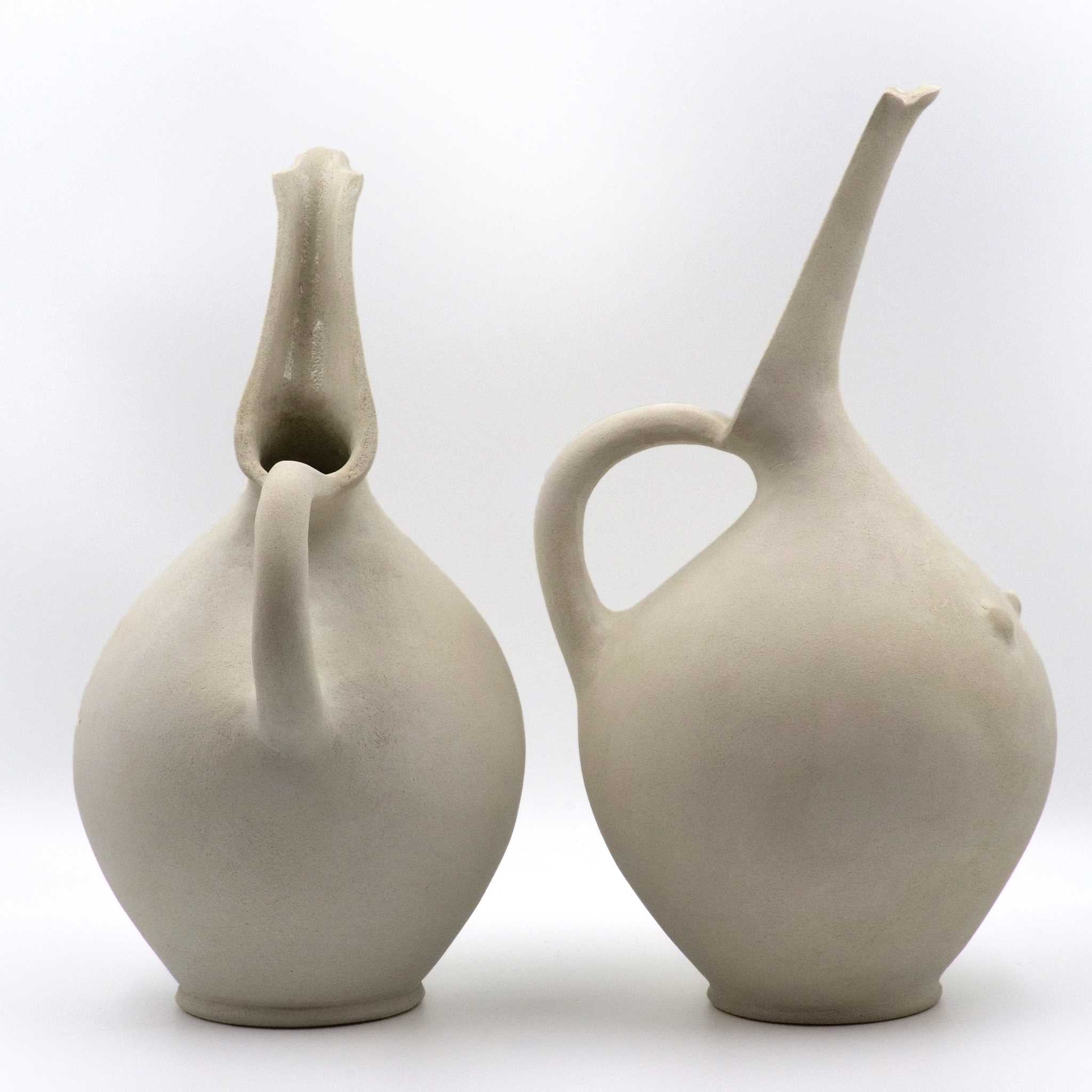Two beige, hand-built ceramic jugs with a subtle breast-like swell and a spout, crafted in glazed stoneware.